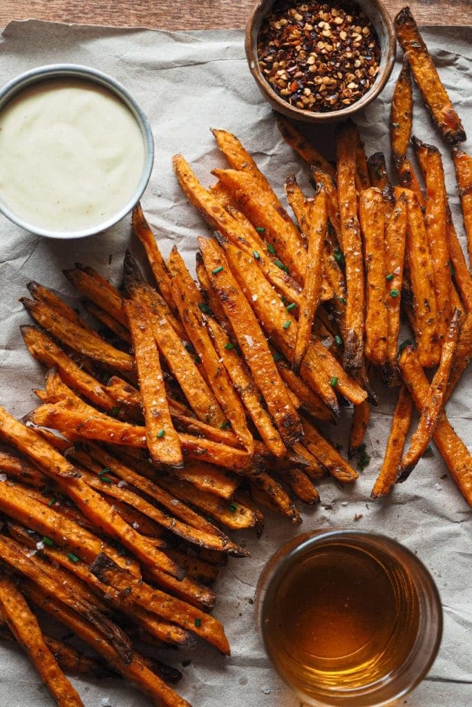 Crispy Baked Sweet Potato Fries with Spicy Mayo, the perfect fall appetizer, side dish, game-day snack, it all works. The cool thing is that they’re healthier than if fried, super easy to make and are actually crispy! There's a small bowl with red pepper flakes at the top, a metallic bowl with the spicy mayo on the left and a glass with beer at the bottom. In the centre and diagonally the sweet potatoes are laying.