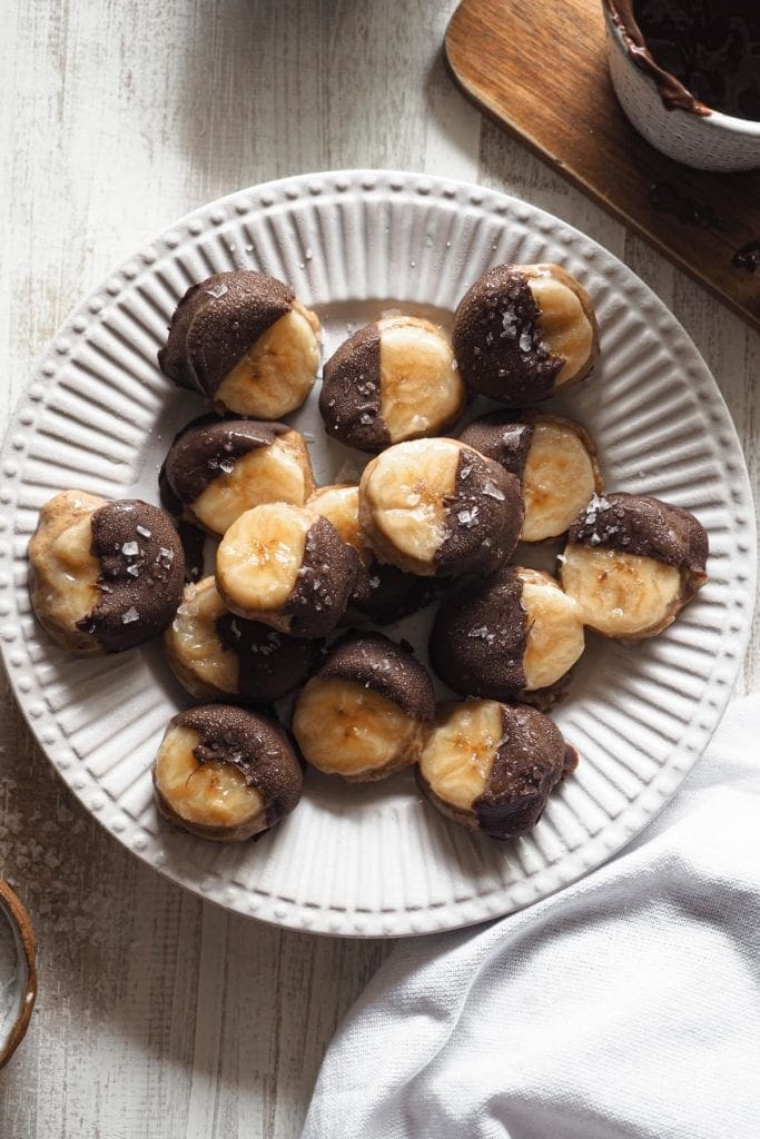 These frosty delicious Banana Almond Butter and Chocolate Bites make the perfect snack. These are the perfect treat! Also gluten free and vegan. Overhead shot of a plate with the bites, salt in a little bowl on the bottom left and a mug with melted chocolate at the top right.