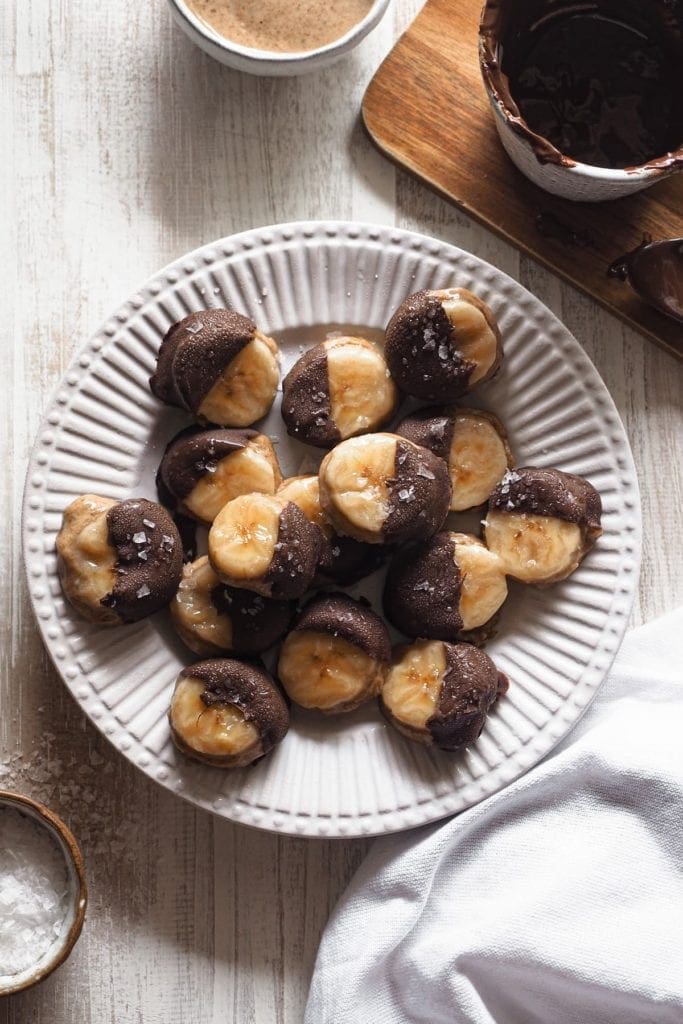 These frosty delicious Banana Almond Butter and Chocolate Bites make the perfect snack. These are the perfect treat! Also gluten free and vegan. Overhead shot of a plate with the bites, salt in a little bowl on the bottom left, a mug with melted chocolate at the top right and a piece of a bowl with almond butter at the top centre.