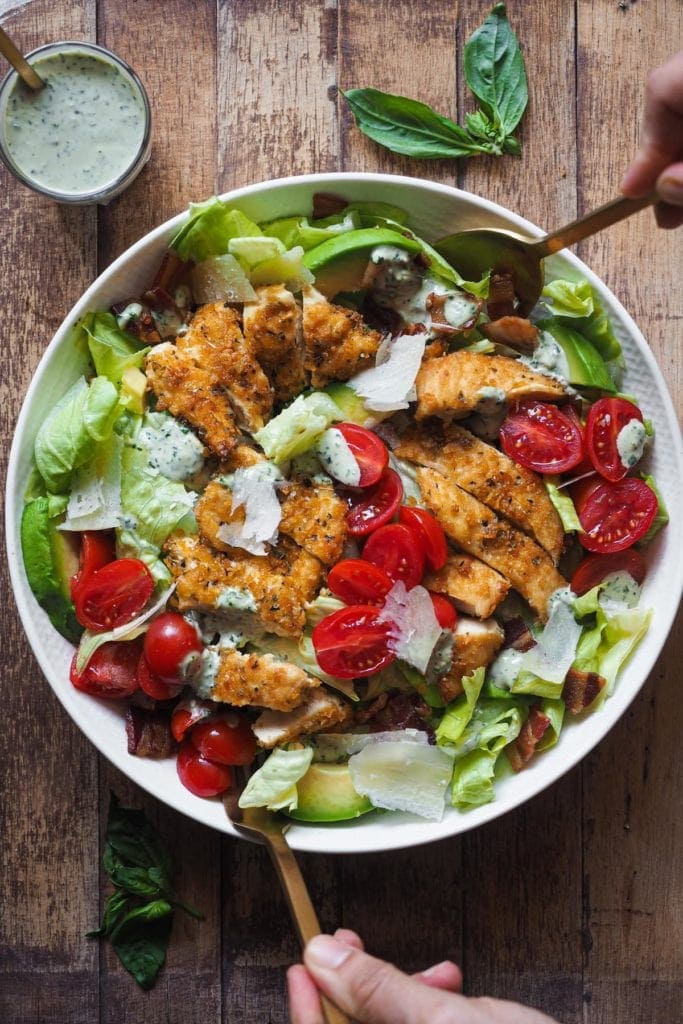 Crispy Chicken and Bacon Salad with Basil Tahini Dressing, an incredible salad that also makes a delicious meal. This salad combo is hard to beat and will make "Salad Meals" something to look forward to. In this picture the salad is served on a plate, placed in the middle, with two hands holding a spoon and a fork as if they were about to toss the salad. On the top left corner there's a small glass with extra dressing and a spoon in it.