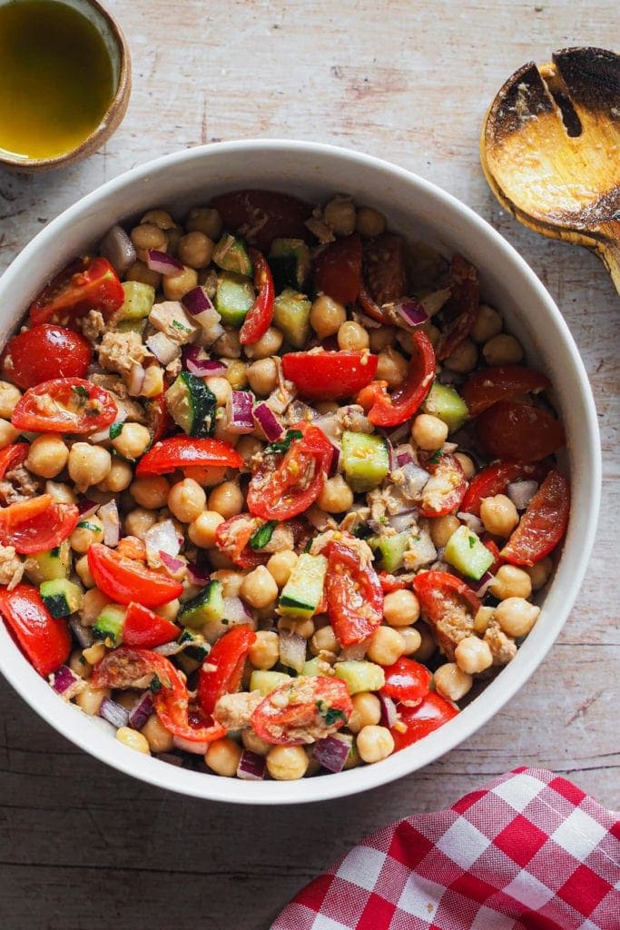 This Mediterranean Chickpea Salad is simple but so flavourful! It combines chickpeas with fresh vegetables and delicious canned tuna, all dressed up with a classic Spanish vinaigrette. It makes a great standalone meal, but it's also perfect as a side or starter. What's more, do you need a packed meal or have a planned picnic or barbecue? This is your salad as it can be prepared in advance and it's so good that everybody will love it. In this overhead shot the salad can be seen served on a bowl with a serving wooden spoon set next to it on the right hand side.