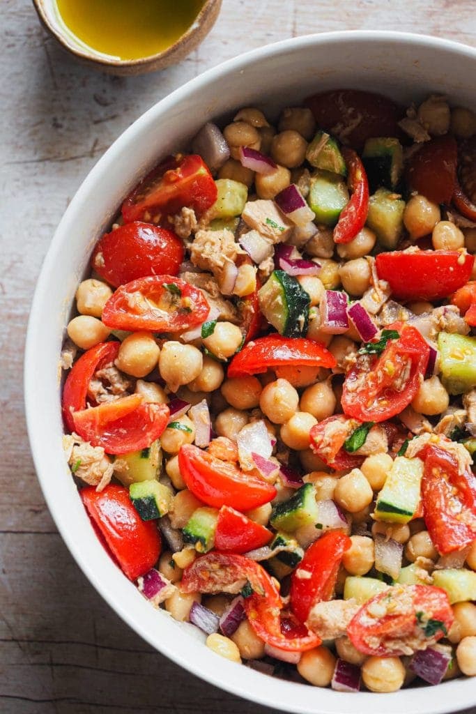 This Mediterranean Chickpea Salad is simple but so flavourful! It combines chickpeas with fresh vegetables and delicious canned tuna, all dressed up with a classic Spanish vinaigrette. It makes a great standalone meal, but it's also perfect as a side or starter. What's more, do you need a packed meal or have a planned picnic or barbecue? This is your salad as it can be prepared in advance and it's so good that everybody will love it. In this overhead shot the salad can be seen served in a bowl with a serving wooden spoon set in it. It is zoomed in, just half of the bowl is shown. At the top left there's a small pinch bowl with extra dressing.