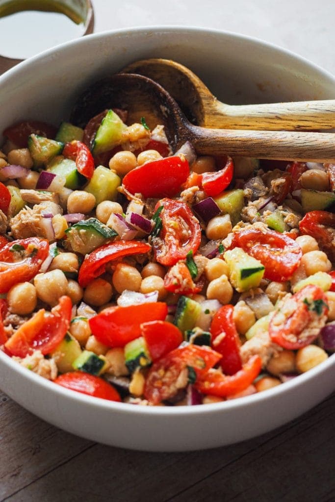 This Mediterranean Chickpea Salad is simple but so flavourful! It combines chickpeas with fresh vegetables and delicious canned tuna, all dressed up with a classic Spanish vinaigrette. It makes a great standalone meal, but it's also perfect as a side or starter. What's more, do you need a packed meal or have a planned picnic or barbecue? This is your salad as it can be prepared in advance and it's so good that everybody will love it. In this 45 angle shot the salad can be seen served in a bowl with a serving wooden spoon set in it.