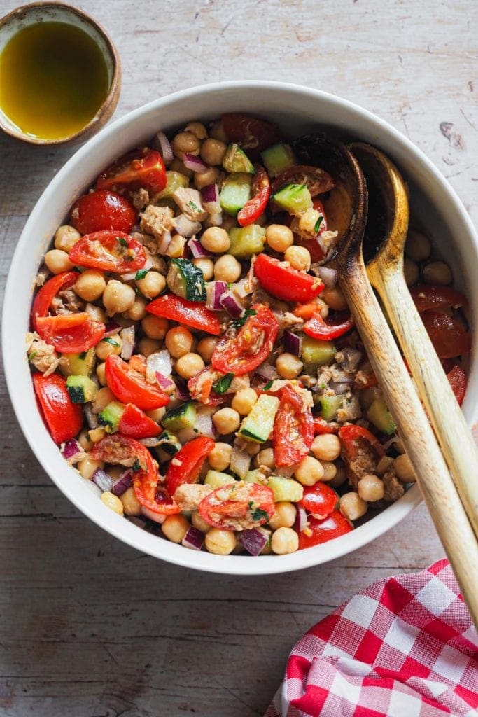 Esta receta de Ensalada Mediterránea de Garbanzos es simple pero muy sabrosa! Combina garbanzos, verduras frescas yatún, todo aderezado con una vinagreta clásica. Es una excelente comida pero lo completa que es, pero también es perfecta como acompañamiento o entrante. Es más, necesitas comida para llevar o tienes planeado un picnic o una barbacoa? Esto es lo que buscas, ya que se puede preparar con antelación y es tan buena que a todo el mundo le va a encantar! En esta foto se ve la ensalada servida en un bol grande con 2 cubiertos de madera para servir apoyados dentro en el lado derecho.