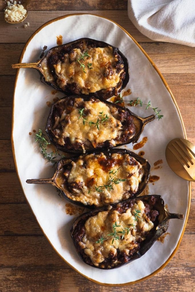 This Sausage Stuffed Aubergine recipe makes a delicious, hearty and wholesome main that everybody will love (no aubergine fans included)! Roasted aubergine is combined with a heavenly tomato sauce, made even better with the addition of sausage and topped with mature cheddar cheese. Overhead shot of the aubergine served on a white plate and garnished with thyme. There is a spoon and a fork leaning on the plate on the right hand side, a kitchen towel on the top right and a tea spoon with cheese at the top left.