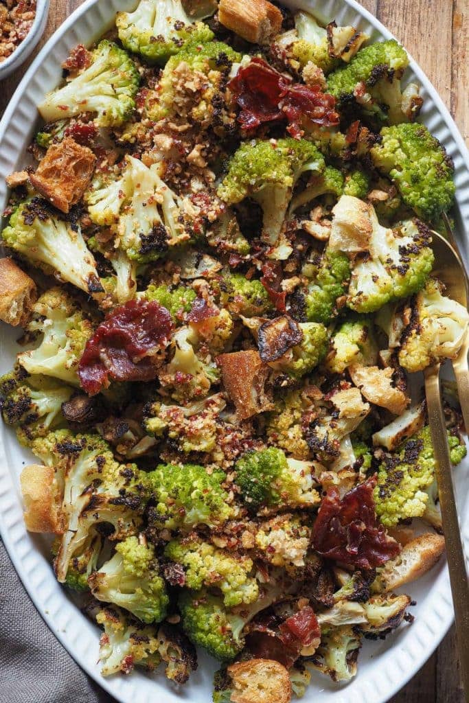 Delicious Roasted Broccoli with Serrano Ham Toasted Breadcrumbs. The roasted broccoli is cooked al dente and gets super crispy on the outside. It's dressed with a toasted breadcrumbs topping that you'll want to add to all your vegetable dishes, or pasta, or... you name it! In this overhead shot, the broccoli can be seen served on an oval white plate, with serving cutlery on the right. The broccoli is topped with the serrano ham toasted breadcrumbs, some pieces of crispy ham and some croutons. The shot is a close up so as to see all the details clearly.