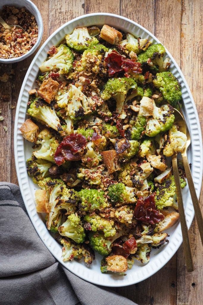 Delicioso Brócoli Asado con Migas de Jamón y Almendra. El brócoli asado queda al dente y súper crujientito por fuera. Lleva un topping de migas que vas a querer añadir a todos tus platos de verduras, o de pasta, o... ¡Lo que quieras! Foto cenital con el brócoli servido en una bandeja blanca ovalada, con las migas por encima, unos trozos de jamón tostado un poco más grande y unos cuantos picatostes. Abajo a la izquierda hay un trapo y arriba a la izquierda un bol pequeño con más migas. Dentro de la bandeja hay cubiertos para servir, a la derecha.