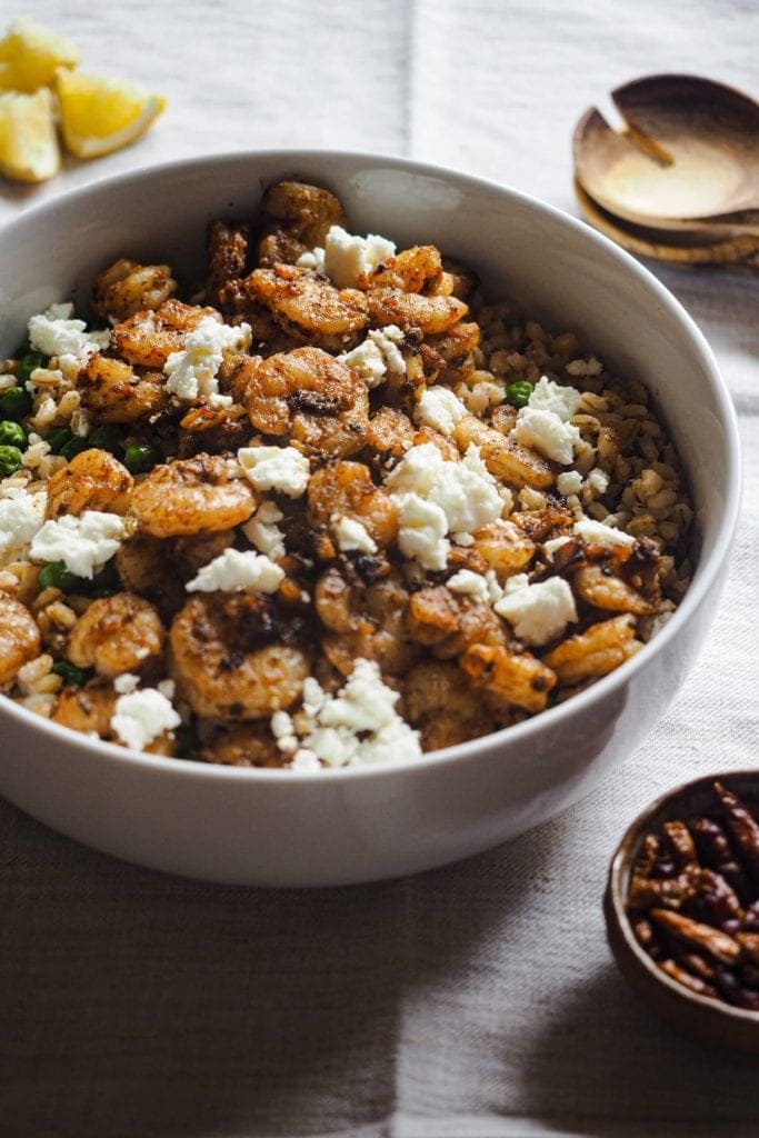 Ensalada Tibia de Gambas y Cebada: súper deliciosa, nutritiva y fácil de preparar. Tiene toques de limón, ajo y pimentón, con guisantes y feta, que se deshace al mezclarlo con las gambas calientes para redondear el plato. En esta foto a 45 grados se ve la ensalada servida en un bol con gajos de limón al fondo a la izquierda, cubiertos de madera para servir la ensalada al fondo a la derecha y un pequeño bol con guindillas delante a la derecha.