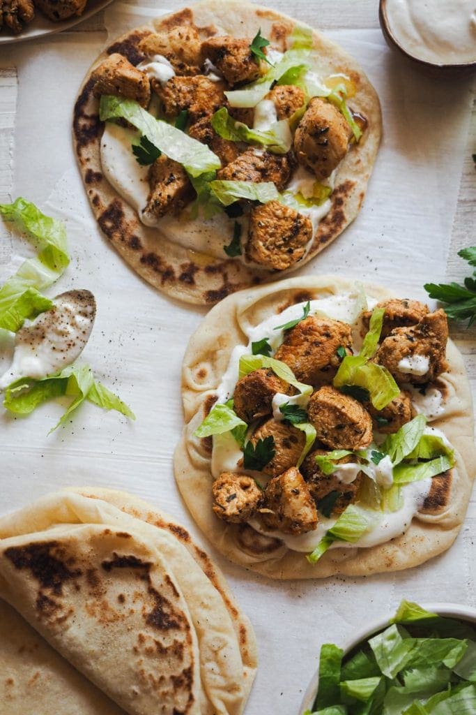 This Mediterranean Spiced Chicken Wraps with Cream Cheese Sauce and Flatbread is so juicy! Chicken marinated with Mediterranean herbs and a spicy and somky touch. Then grilled and served with a delicious cream cheese sauce and Turkish flatbread. It makes a great meal served like a wrap or pita. This is an overhead shot of two flatbreads layered with cream cheese sauce, some spiced chicken, the lettuce and parsley. There is a folded flatbread on the bottom left corner and some lettuce on the bottom right.