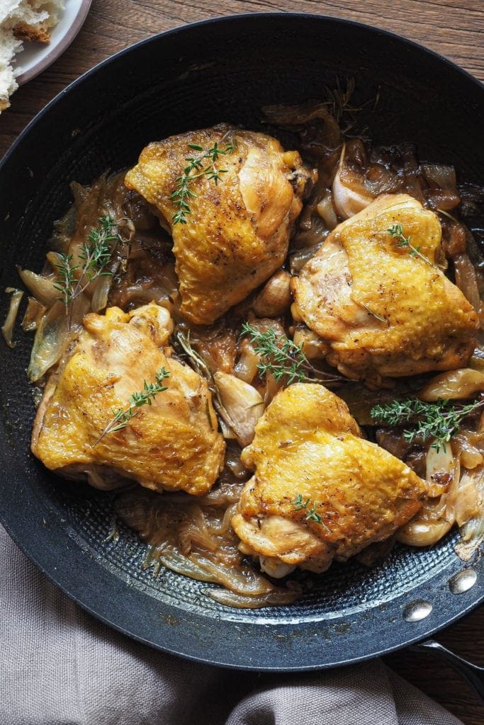 Jugoso Pollo al Cava (o Pollo al Champagne), sencillo, lleno de sabor y te hace quedar genial si tienes una comida con amigos o familia! Lleva cebolla y ajo, que quedan a punto de caramelo, y hierbas aromáticas para añadir un toque más de sabor. Es perfecto para hacer en modo batchcooking y así tener para más de un día. Y bueno, qué decir de las recetas de la abuela... ¡nunca fallan! Aquí el pollo se ve de cerca, servido en una sartén profunda de hierro, con pan a un lado y un trapo en otro. La foto está centrada.