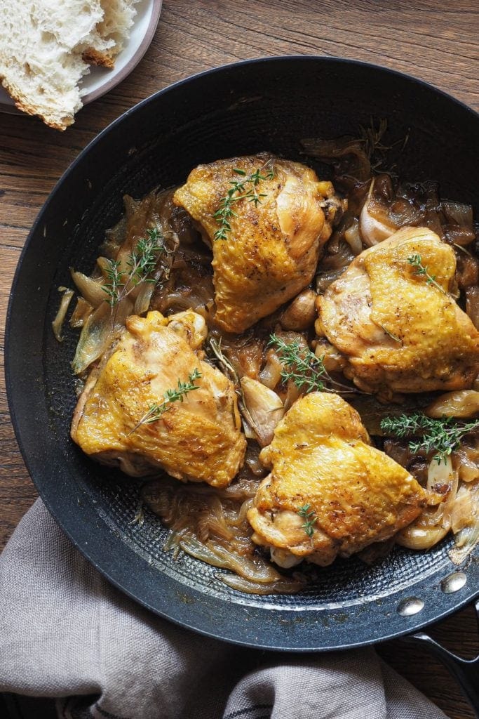 Jugoso Pollo al Cava (o Pollo al Champagne), sencillo, lleno de sabor y te hace quedar genial si tienes una comida con amigos o familia! Lleva cebolla y ajo, que quedan a punto de caramelo, y hierbas aromáticas para añadir un toque más de sabor. Es perfecto para hacer en modo batchcooking y así tener para más de un día. Y bueno, qué decir de las recetas de la abuela... ¡nunca fallan! Aquí el pollo se ve de cerca, servido en una sartén profunda de hierro, con pan a un lado y un trapo en otro. La sartén está recortada por la derecha en la foto.