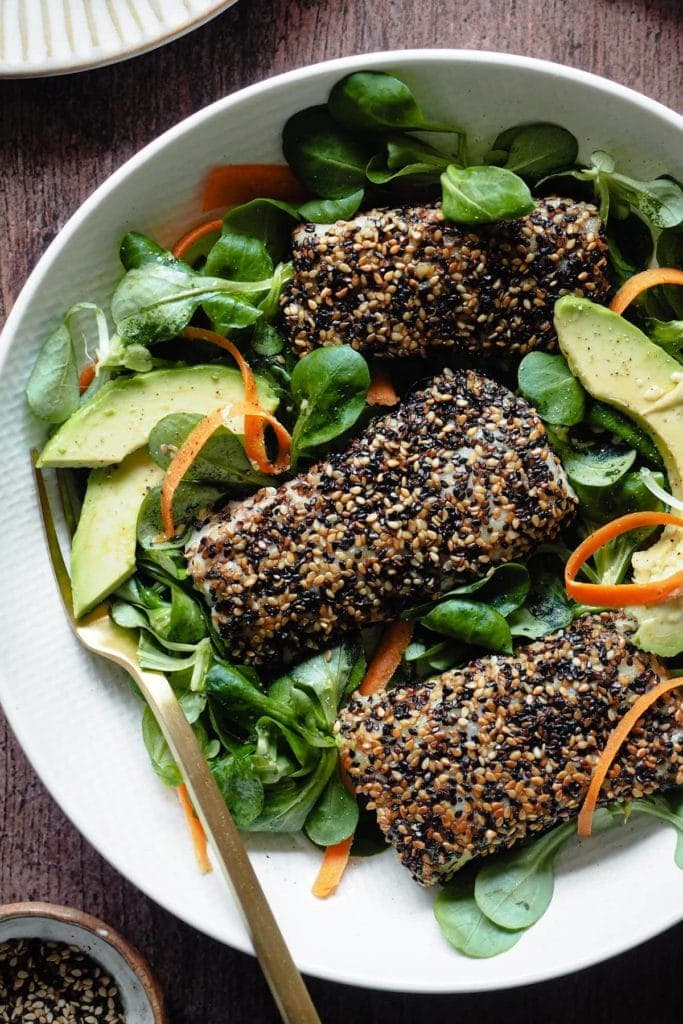 Delicious Sesame-Crusted Hake. It takes 10 minutes max to put together and will make the whole family fall in love with fish! Serving it with a bright salad also helps and the lemony mustard vinaigrette just pairs so well with fish. In this picture 3 sesame-crusted hake fillets are placed on top of an avocado, carrot and lamb's lettuce salad. There's a fork in the plate too and pinch bowl with sesame seeds at the bottom left corner of the picture.