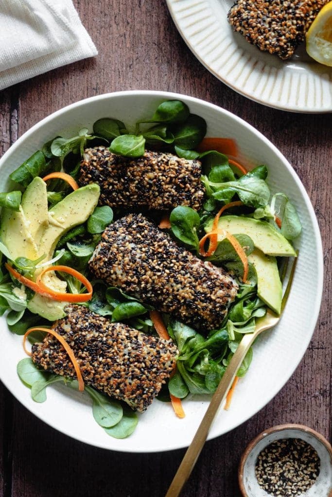 Delicious Sesame-Crusted Hake. It takes 10 minutes max to put together and will make the whole family fall in love with fish! Serving it with a bright salad also helps and the lemony mustard vinaigrette just pairs so well with fish. In this picture 3 sesame-crusted hake fillets are placed on top of an avocado, carrot and lamb's lettuce salad. There's a fork in the plate too and pinch bowl with sesame seeds at the bottom right corner of the picture.