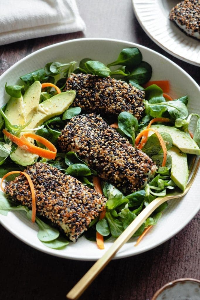 Delicious Sesame-Crusted Hake. It takes 10 minutes max to put together and will make the whole family fall in love with fish! Serving it with a bright salad also helps and the lemony mustard vinaigrette just pairs so well with fish. In this picture 3 sesame-crusted hake fillets are placed on top of an avocado, carrot and lamb's lettuce salad. There's a fork in the plate too on the right side.