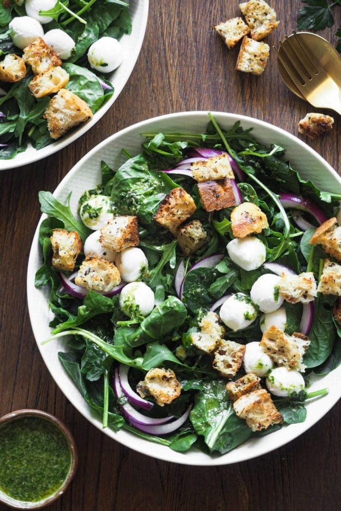 Esta ensalada de espinacas con vinagreta de alcaparras combina todos los ingredientes clave de una buena ensalada: verde del bueno con espinacas y rúcula, crujiente con picatostes, color con el contraste blanco de la mozzarella y el morado de la cebolla, todo ligado con una deliciosa vinagreta de alcaparras. Si tienes picatostes a mano, en 5 minutos la tienes lista! Foto cenital del plato de ensalada. Este está en el centro y arriba a la izquierda se aprecia un trocito de otro plato con más ensalada. Arriba a la derecha hay una cuchara y tenedor dorados rodeados por picatostes. Abajo a la derecha hay un pequeño bol con aliño extra.