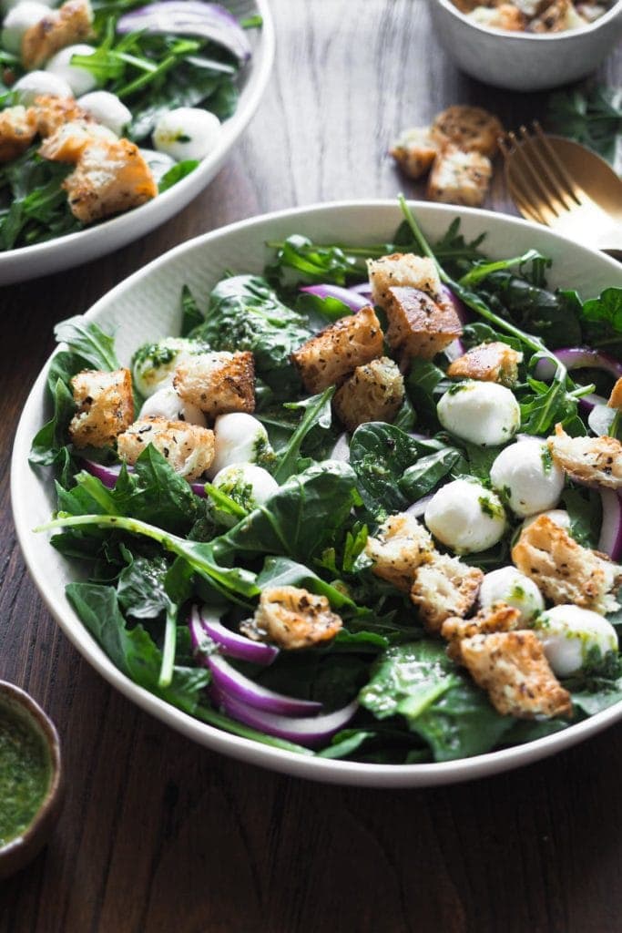 Esta ensalada de espinacas con vinagreta de alcaparras combina todos los ingredientes clave de una buena ensalada: verde del bueno con espinacas y rúcula, crujiente con picatostes, color con el contraste blanco de la mozzarella y el morado de la cebolla, todo ligado con una deliciosa vinagreta de alcaparras. Si tienes picatostes a mano, en 5 minutos la tienes lista! Foto a 45 grados del plato de ensalada. Este está en el centro y arriba a la izquierda se aprecia un trocito de otro plato con más ensalada. Arriba a la derecha hay una cuchara y tenedor dorados rodeados por picatostes y perejil. Abajo a la derecha hay un pequeño bol con aliño extra.