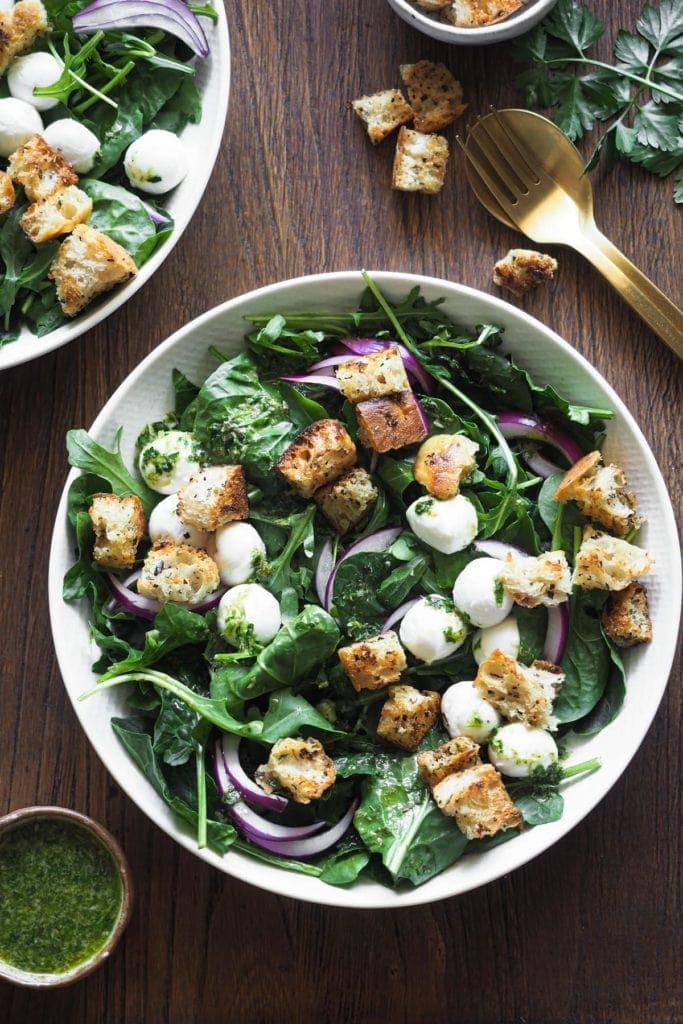 This salad combines all the things that are key in a salad: great greens, crunchy croutons for good texture, colour notes with the white mozzarella and red onions, plus a delicious punchy capers vinaigrette to dress it all up. If you already have homemade croutons in hand, it just takes 5 minutes to put together! overhead shot of a plate with the salad. There's a bit of another salad plate on the top left corner, a golden fork and spoon on the top right surrounded with croutons and parsley and a small bowl with extra dressing on the bottom left corner.