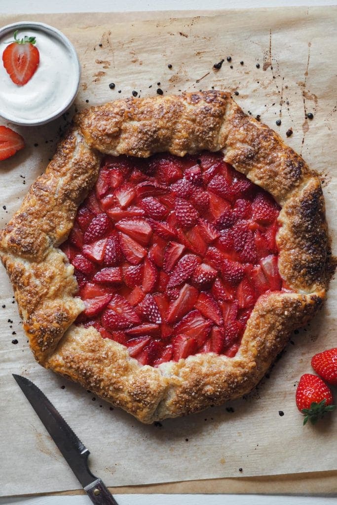 Bursting with fresh spring strawberries this Strawberry Galette with Vanilla Whipped Cream on the side is truly a spring and summer treat. Made with a quick flaky pastry and accompanied with vanilla whipped cream, it's a twist on the classic strawberries and cream dessert that will make everybody WOW. It's delicious, simple and super gorgeous. You can’t go wrong with this one, I promise! Here we can see an overhead shot of the whole galette. At the top left corner there is a small bowl with the whipped cream with half strawberry on top and half strawberry on its left side. On the bottom right, next to the galette, there are 2 strawberries. On the bottom left corner there's a serrated knife.