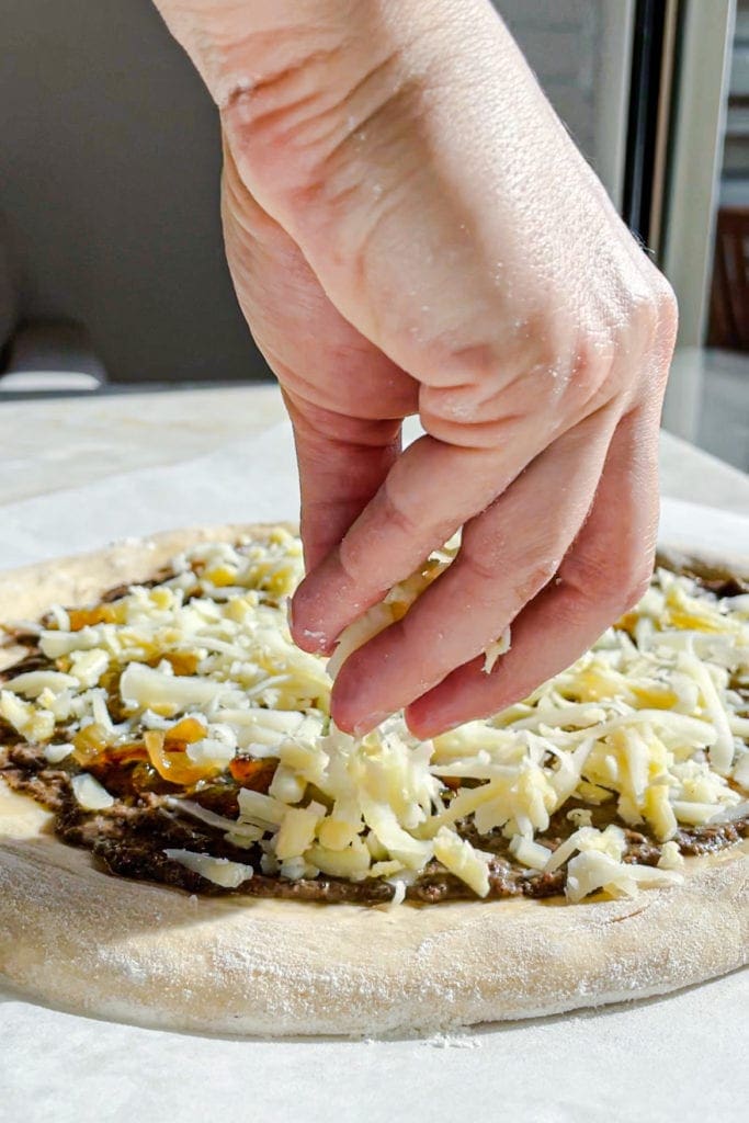 Pizza de Patata y Burrata con Trufa antes de hornearla, poniendo queso cheddar rallado sobre la base de trufa y cebolla caramelizada. ¡Para morirse de buena! | Receta de Paula's Apron