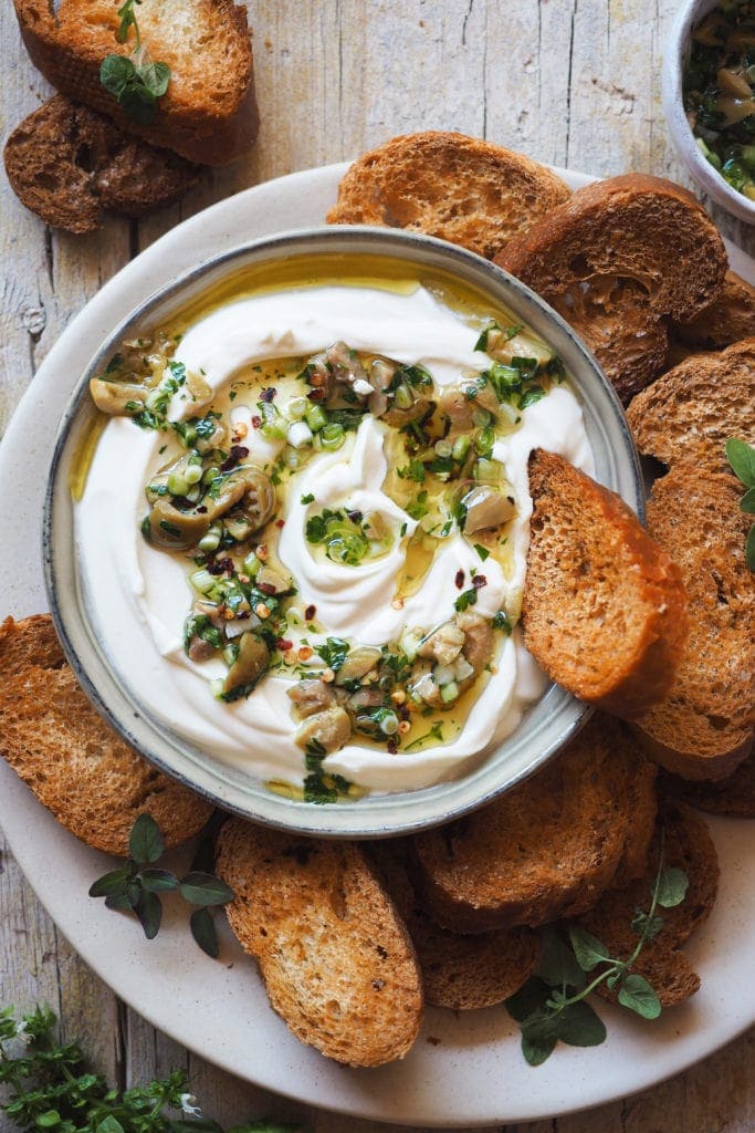 This Garlic Confit Goat Cheese Dip with Spicy Lemony Olives is creamy, tasty and easy to make. Oh! And it's a 3 in one dip: a spreadable garlic confit cheese base, homemade toasts and the most delicious olive topping. It makes a delicious appetiser and it's perfect for any occasion and all year round! | Paula's Apron Recipe