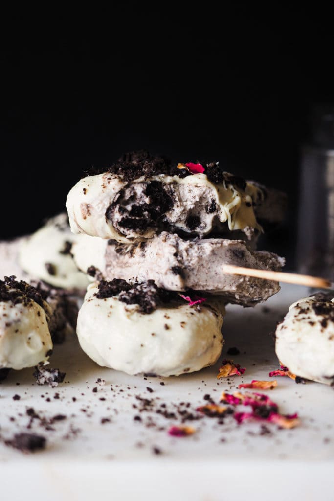 Helado Cremoso de Oreo con Solo 3 Ingredientes, perfecto para combatir el calor y matar antojos de dulce. La cobertura de chocolate blanco y oreo es totalmente opcional. Así que si quieres evitarte un par de pasos e ingredientes, puedes congelar el helado en un recipiente hermético o molde directamente, y es que tal cual ya está buenísimo. En esta foto frontal, los helados están apilados y el último tiene un mordisco. Se ve el interior cremoso con trocitos de oreo.| Receta de Paula's Apron