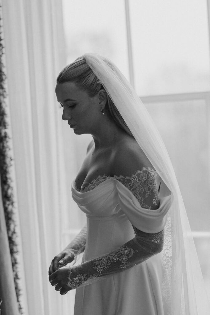 A bride in an off-the-shoulder lace wedding dress and veil stands indoors near a window, looking down thoughtfully with her hands together. Captured in black and white by a creative wedding photographer.