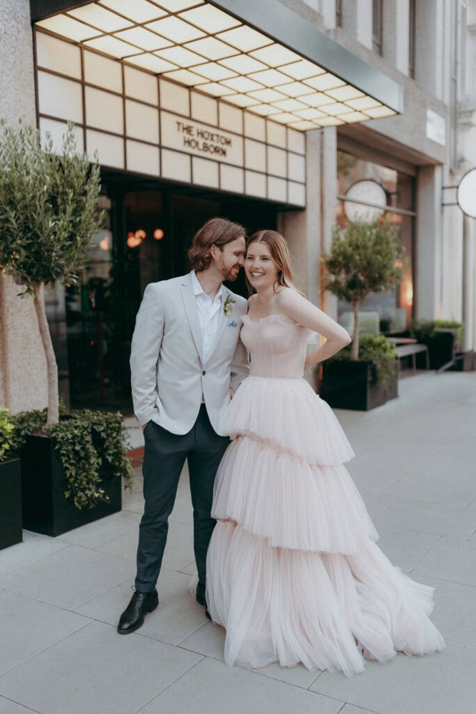 A smiling couple poses outside a modern building, captured beautifully by a wedding photographer. The woman wears a tiered, light pink strapless gown, while the man in a light gray suit and white shirt leans close, both looking happy and relaxed.