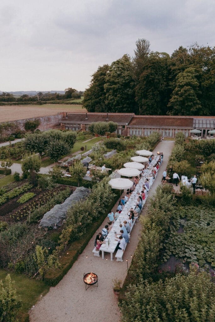 Aerial view of a long outdoor dining table set for a gathering in a lush garden, captured by a creative wedding photographer, with people seated under white umbrellas, surrounded by greenery and trees beneath a cloudy sky.