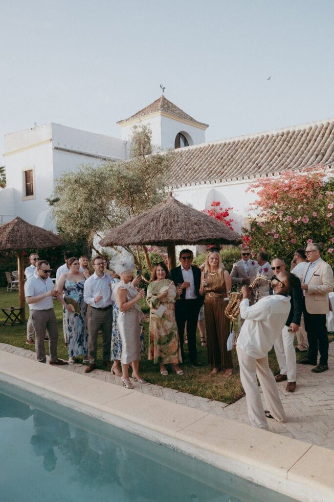A group of people dressed in semi-formal attire gather beside a pool at an outdoor event, smiling and watching two musicians perform, as a Creative Wedding Photographer captures the scene. White buildings and flowering greenery frame the clear sky backdrop.