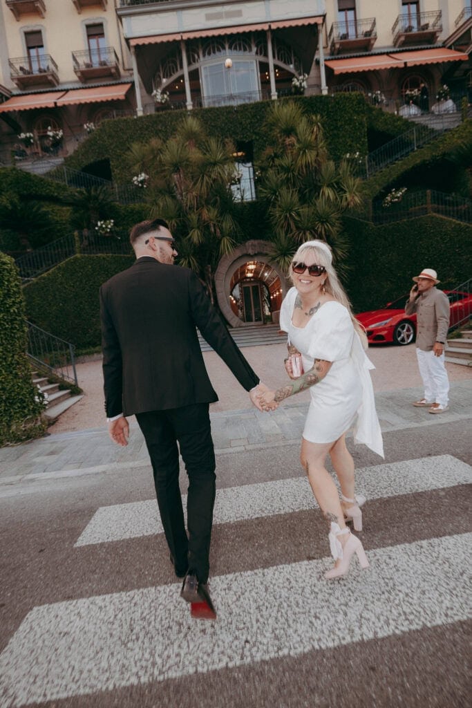 A couple holding hands crosses a street towards a lush, grand hotel. Captured by a creative wedding photographer, the woman in a white dress smiles back in sunglasses, while the man in a dark suit looks ahead. A red car and another person are visible behind them.