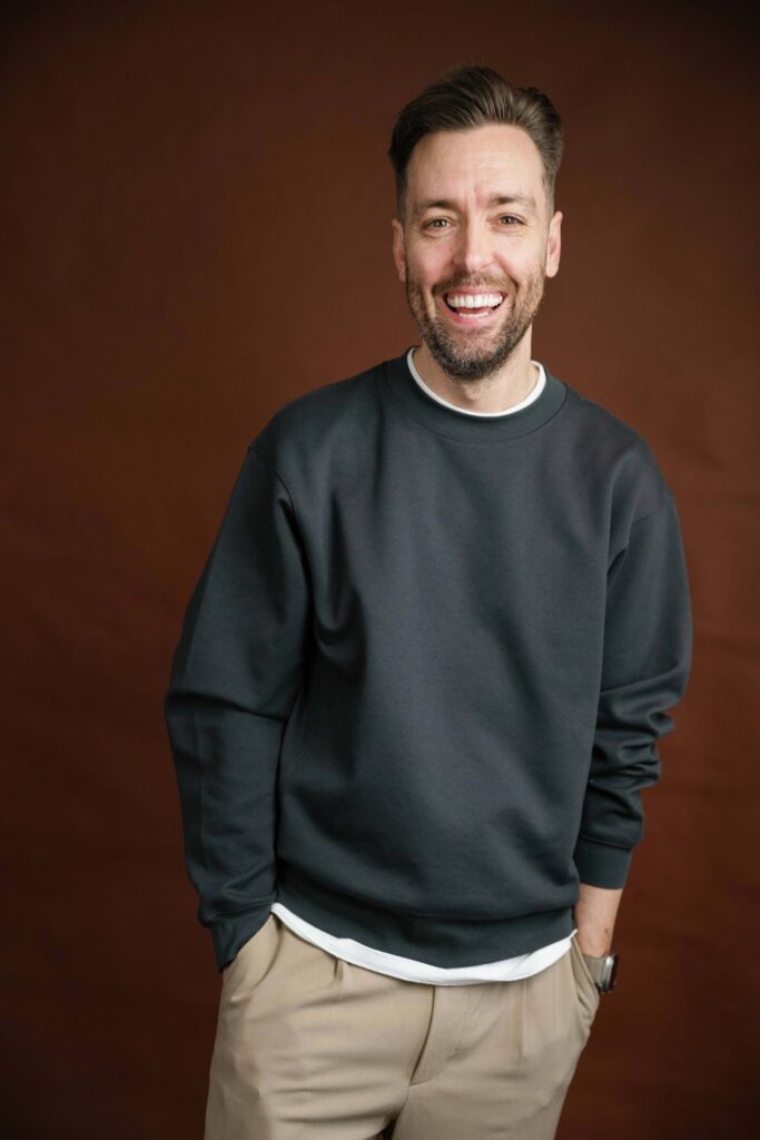 A man with short brown hair and a beard smiles at the camera. He is wearing a dark gray sweatshirt, beige pants, and a watch, standing against a brown background with one hand in his pocket.