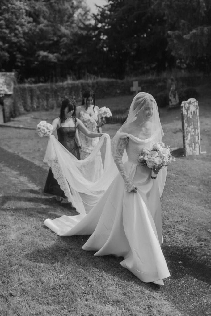 A bride in a long, elegant gown and veil walks outdoors, holding a bouquet, while two bridesmaids behind her hold her train. Captured by a Creative Wedding Photographer, the black and white shot features gravestones and trees in the grassy background.