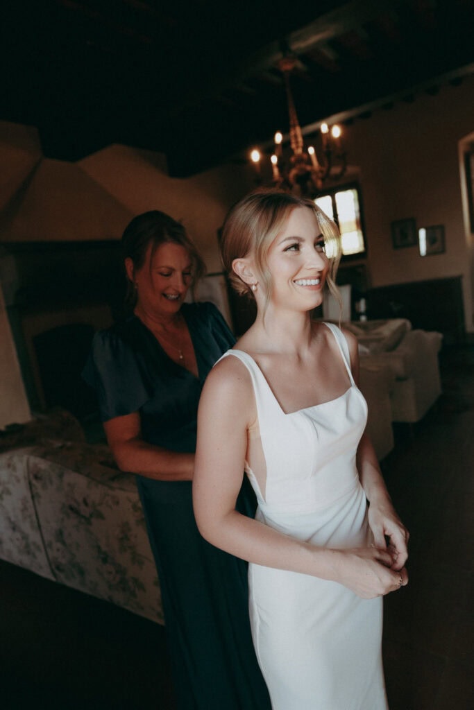 A smiling bride in a white dress stands indoors at Borgo di Pietrafitta as another woman helps fasten her dress. The warmly lit room features rustic decor, with a chandelier and large window in the background.