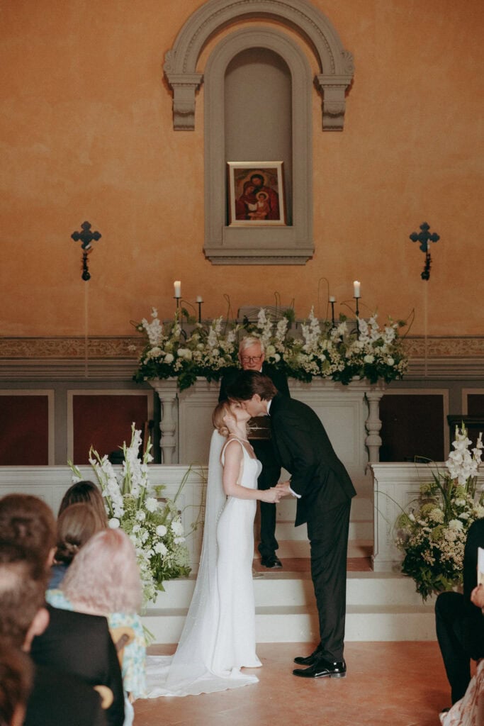 A bride and groom share a kiss at the altar during their wedding ceremony at Borgo di Pietrafitta, with a priest behind them and white floral arrangements and candles adorning the altar. Guests are seated in the foreground.