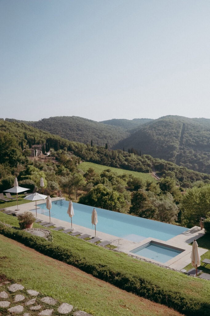 An infinity pool at Borgo di Pietrafitta with closed umbrellas and lounge chairs overlooks rolling green hills and lush countryside under a clear sky. Stone paths and manicured grass surround the peaceful pool area.