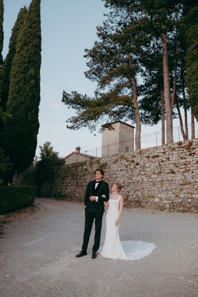 A bride in a white gown stands beside a groom in a black suit on a gravel path at Borgo di Pietrafitta, with tall trees and a stone wall in the background under a clear sky.