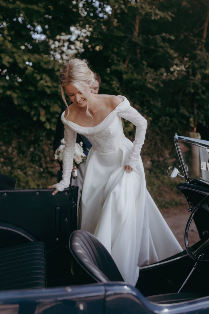 A bride in an off-the-shoulder white wedding gown smiles as she steps into a vintage car. Lush green foliage surrounds her, capturing a romantic outdoor moment perfect for any Cotswolds Wedding Photographer.