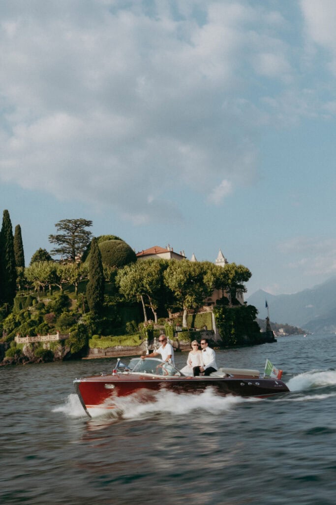Three people ride in a vintage wooden motorboat on a lake, with lush greenery and a villa on a hillside in the background under a partly cloudy sky.