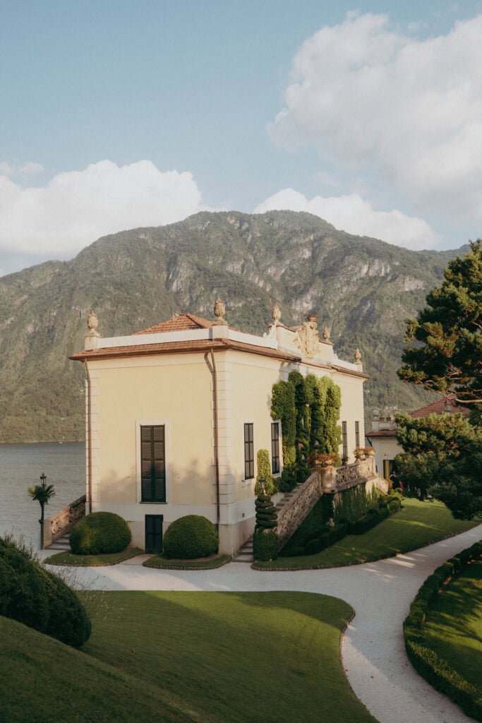 A yellow villa with tall windows and manicured greenery sits by a lake, backed by tree-covered mountains and a partly cloudy sky. Curved pathways and trimmed lawns surround the building.