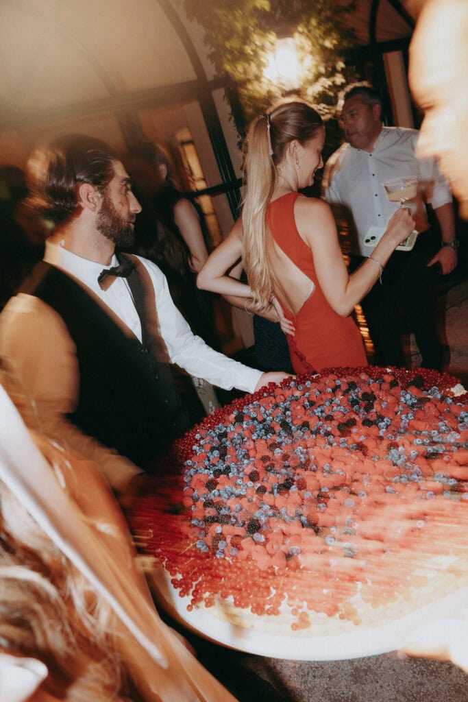 A formally dressed waiter carries a large, round berry tart at a lively indoor event; guests in elegant attire, including a woman in a red dress, mingle and hold drinks in the warmly lit atmosphere.