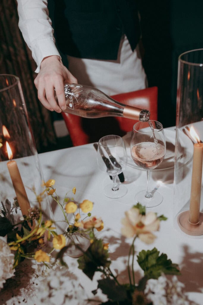 A London Wedding Photographer captures a person pouring rosé wine into a glass on an elegantly set table adorned with candles, flowers, and sparkling glassware.