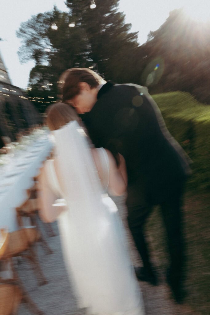 A blurred photo shows a bride in a white dress and veil walking beside a person in a black suit outdoors at Borgo di Pietrafitta, near a long table with chairs. Sunlight and greenery are visible in the background.