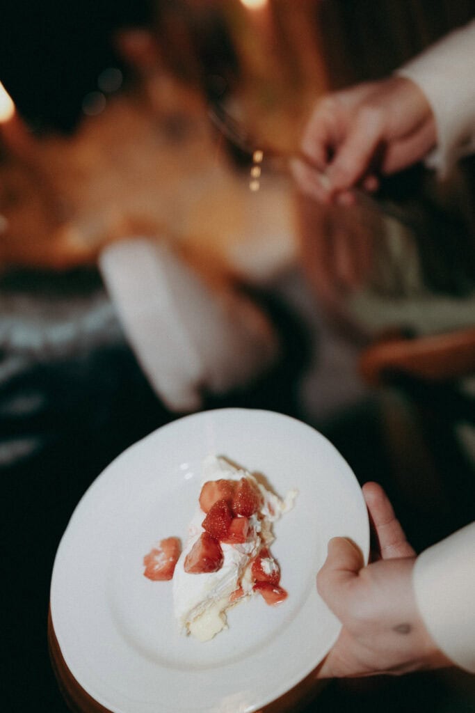 A person holds a white plate with a slice of cake topped with whipped cream and strawberries at Borgo di Pietrafitta. The other hand holds a fork, about to take a bite. The softly blurred background evokes rustic charm.