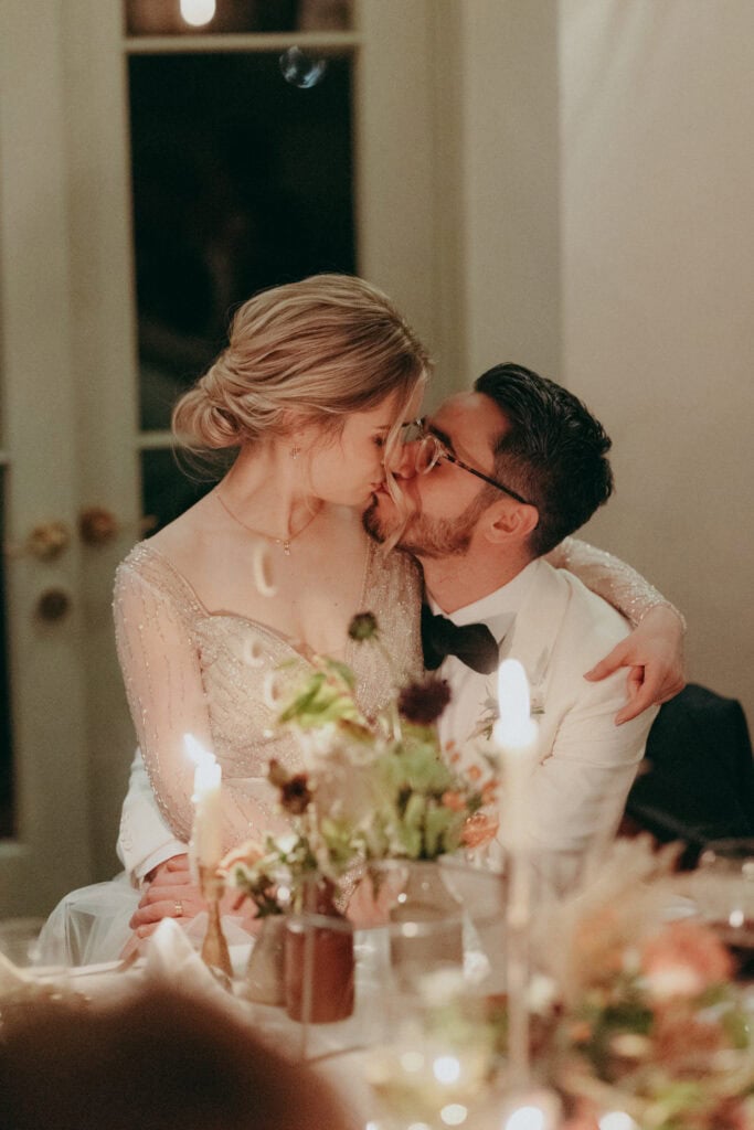 A bride and groom share a romantic kiss while seated at a candlelit table, surrounded by floral arrangements. Captured by a London Wedding Photographer, the sparkling gown and white tuxedo jacket create an intimate and warm setting.
