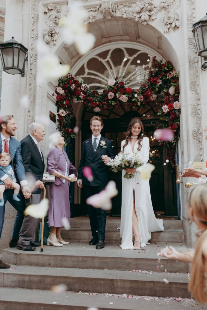 A bride and groom smile as they exit a flower-adorned venue, surrounded by family. Captured by a London Wedding Photographer, joyful guests toss petals in celebration while a man on the left holds a child.