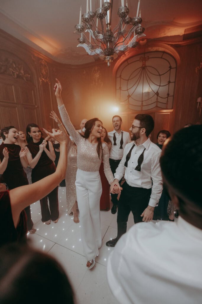 A joyful woman in white pants and a sparkly top dances and raises her arm while holding hands with a smiling man in a tuxedo shirt, captured by a London Wedding Photographer at a lively indoor celebration with friends.