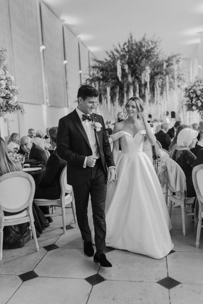 A bride and groom walk together inside a decorated wedding venue, smiling and holding hands. Captured by a London Wedding Photographer, guests are seated at tables while floral arrangements hang from the ceiling in this black-and-white scene.