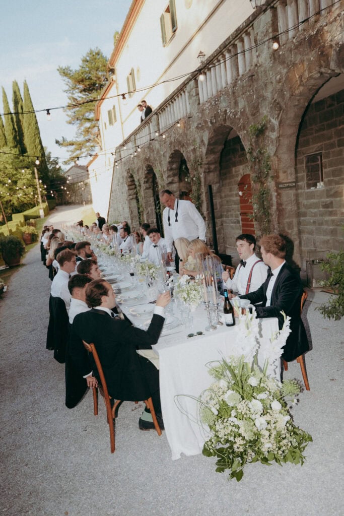 A long outdoor dining table is set for a formal event beside a stone building at Borgo di Pietrafitta. Guests in suits and white shirts are seated, one stands to speak. White flowers and string lights elegantly decorate the scene.