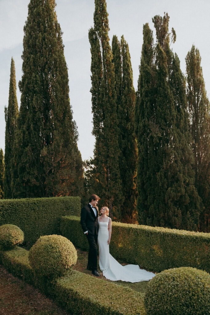 A bride and groom stand together in the manicured gardens of Borgo di Pietrafitta, surrounded by tall cypress trees and neatly trimmed hedges, sharing a quiet moment on their wedding day.