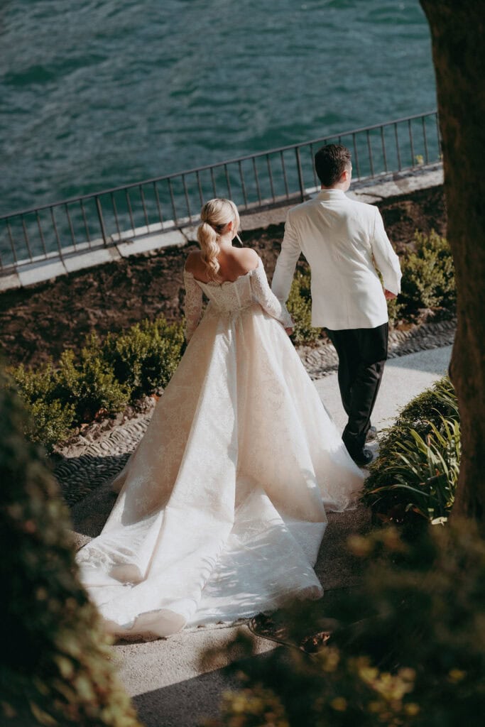 A bride in an elegant white gown walks hand in hand with a groom in a white jacket by a waterfront, with sunlight highlighting the scene and greenery surrounding the path.